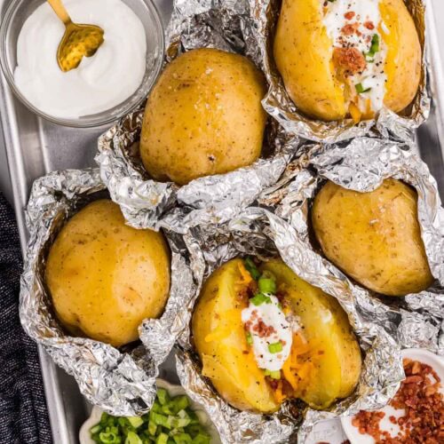 cooking sheet with opened tin foiled potatoes and containers of green onion, bacon bits and sour cream with spoon, some potatoes halved and dressed with sour cream, bacon bits, and green onions