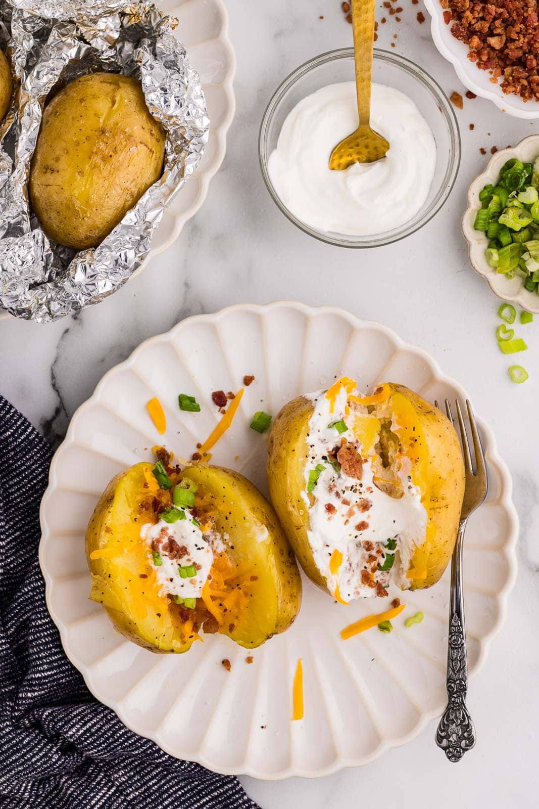 overhead view of white dinner plate with two halved potatoes dressed with sour cream, bacon bits and green onion, surrounded by bowls of tin foiled potatoes, bowl with sour cream, bowl of bacon bits, and bowl of green onions
