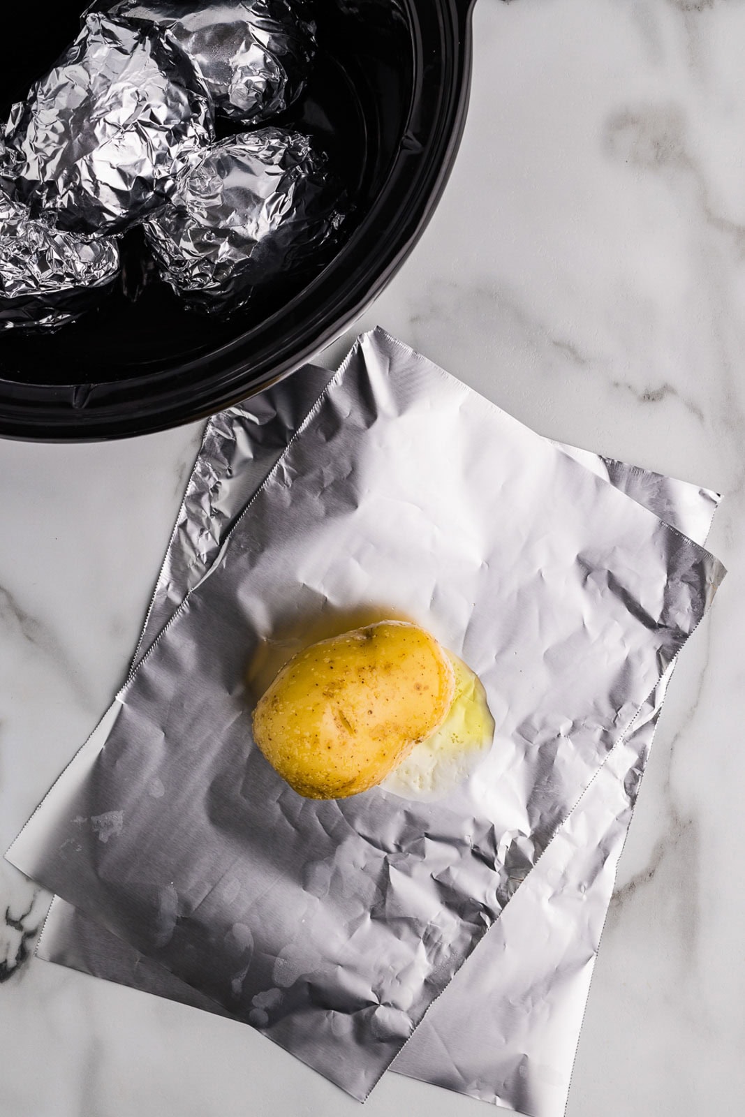sheets of tinfoil with one potato on top with black slow cooker containing prepared potatoes in background