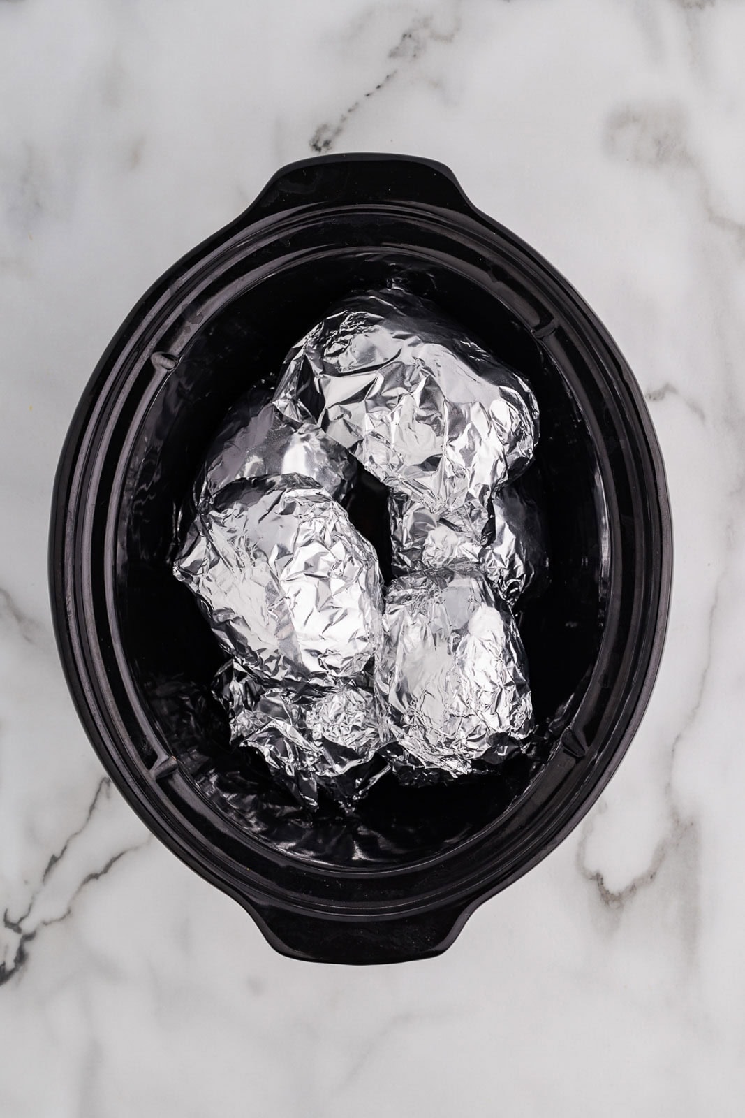 overhead view of black slow cooker containing six tin foiled potatoes