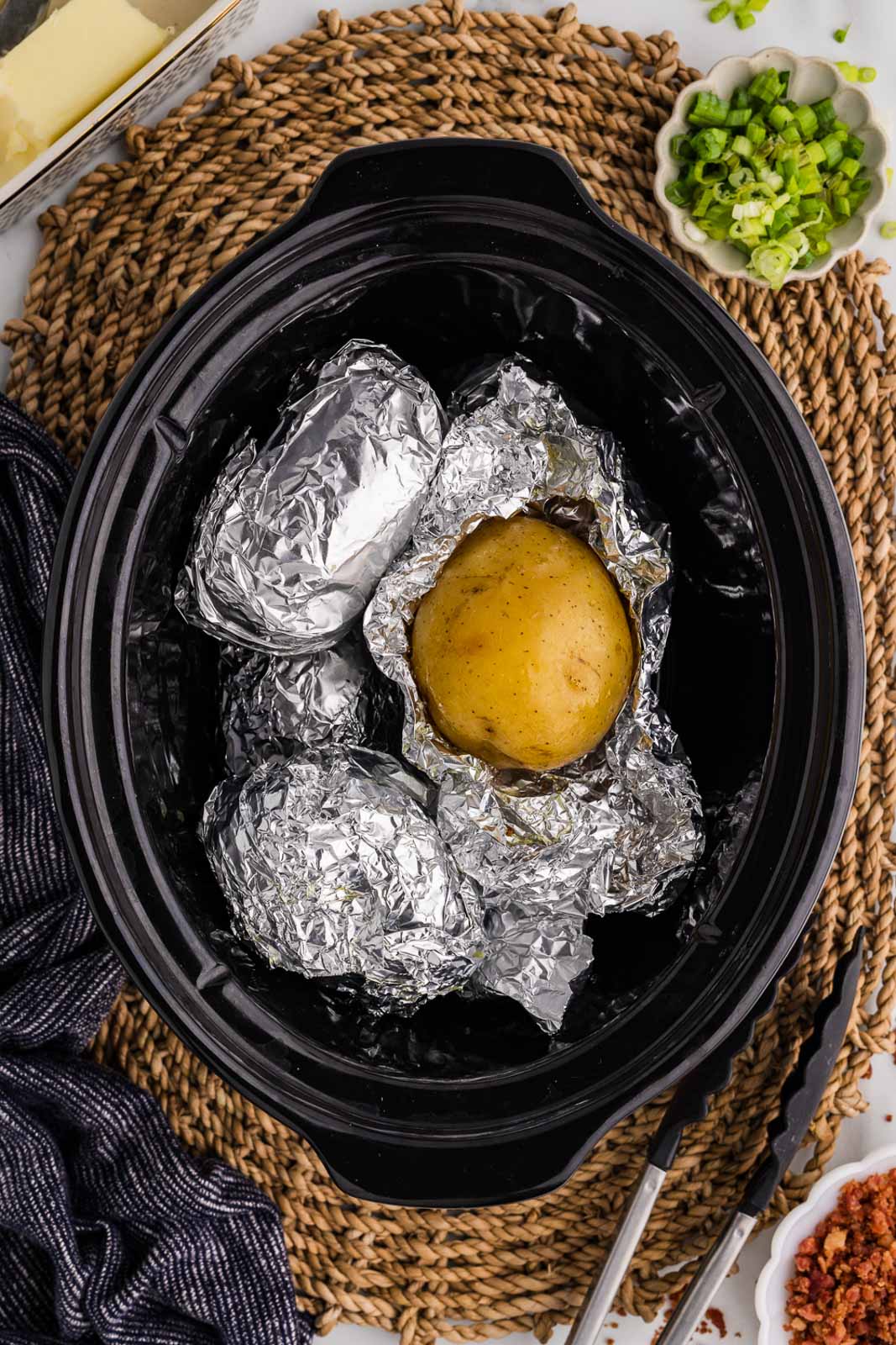 overhead view of black slow cooker with tin foiled potatoes, one uncovered with butter, green onion, and bacon bits on side