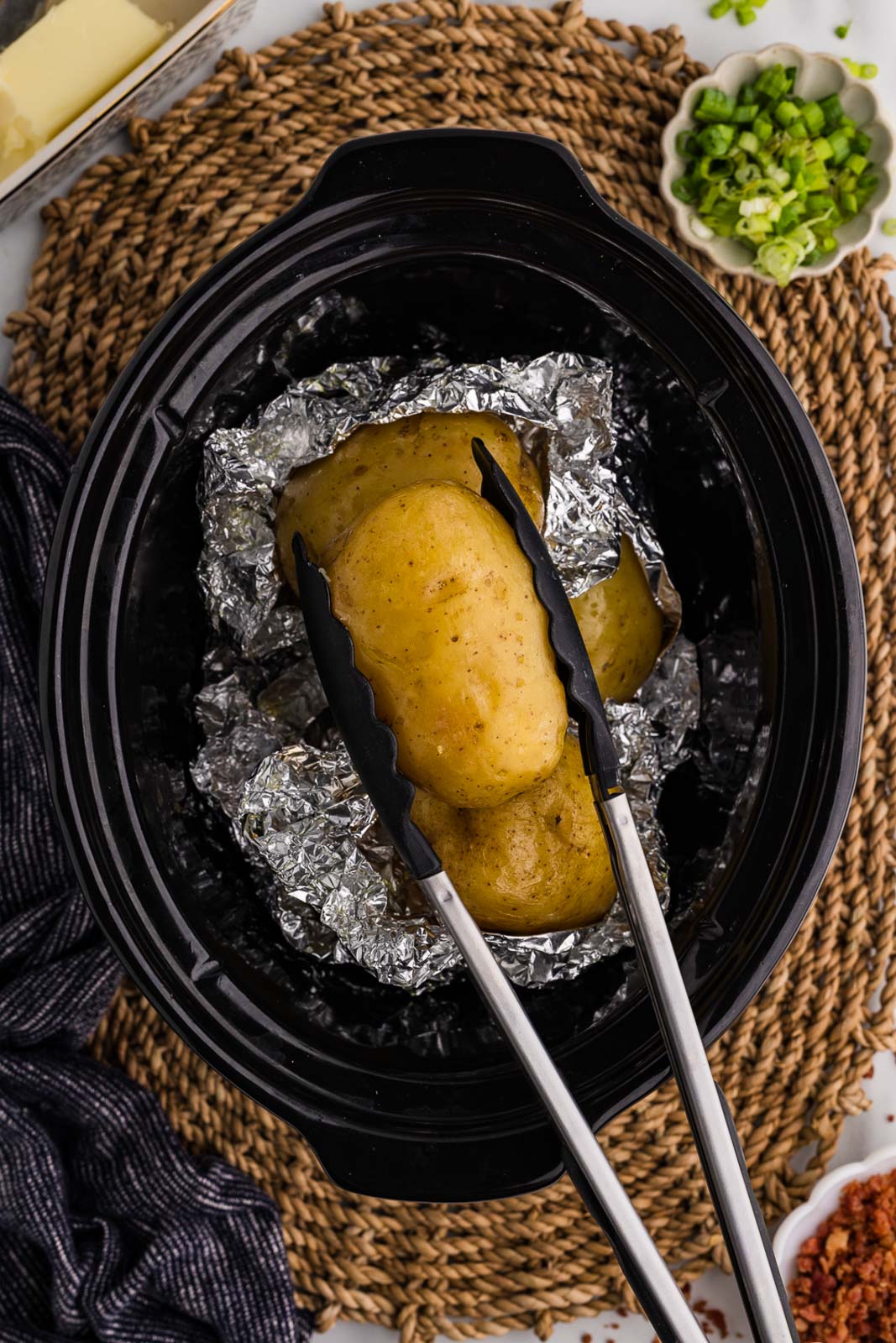overhead view of black slow cooker with tin foiled potatoes, several uncovered, one being held by tongs, surrounded by green onions, butter and bacon bits