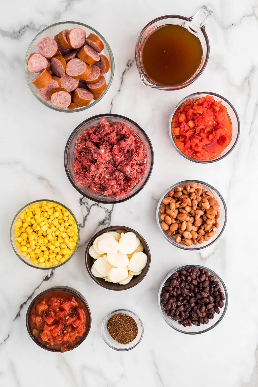 ingredients to include bowls of smoked sausage, ground beef, pinto beans, black beans, corn, sliced potatoes, diced tomatoes, diced tomatoes with chilis, chili seasoning, and a measuring cup of beef broth