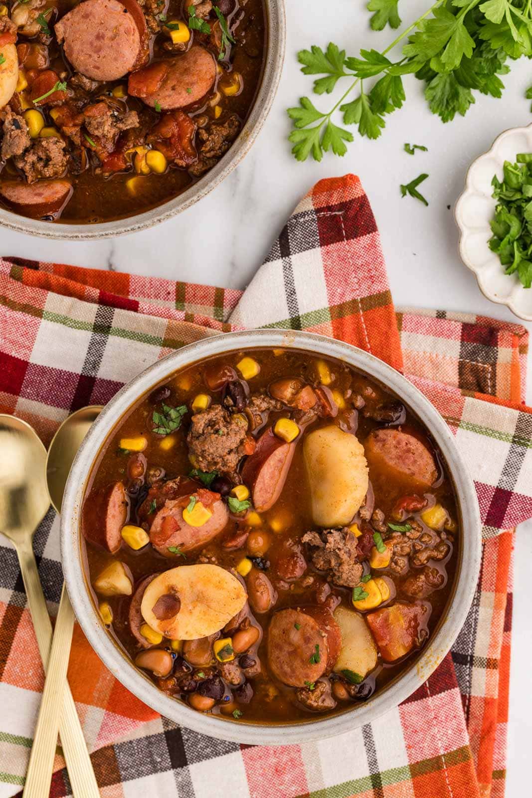 overhead view of bowls of cowboy stew on checkered napkin, two gold spoons and dish of parsley