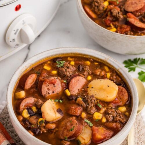 overhead view of two bowls of cowboy stew, one on a tan placemat, one gold spoon and white slow cooker in the background