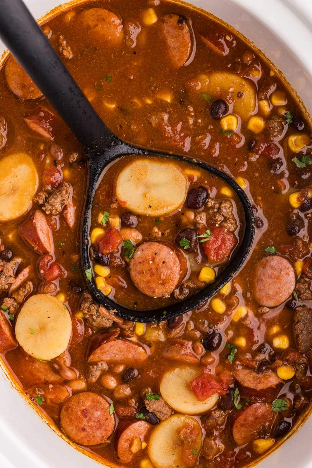 overhead closeup of white slow cooker of cowboy stew with black ladle