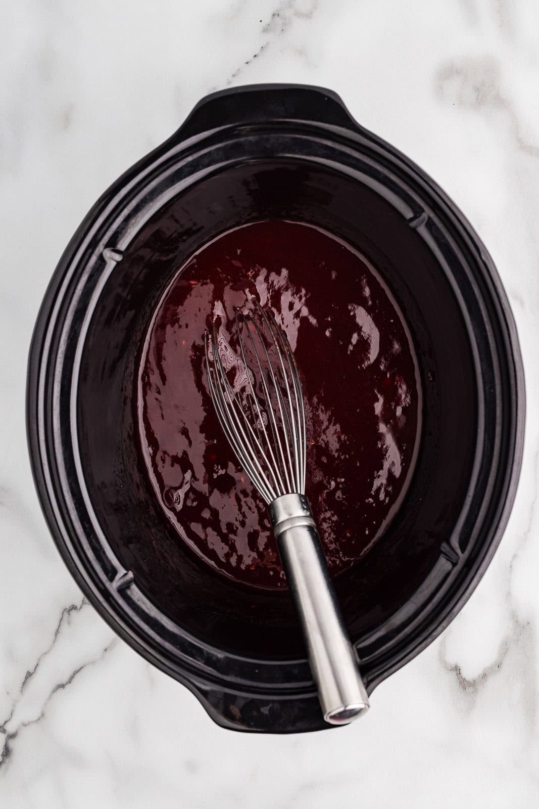 overhead view of black slow cooker containing cranberry sauce, ketchup, maple syrup, orange juice, vinegar and red pepper flakes with silver whisk