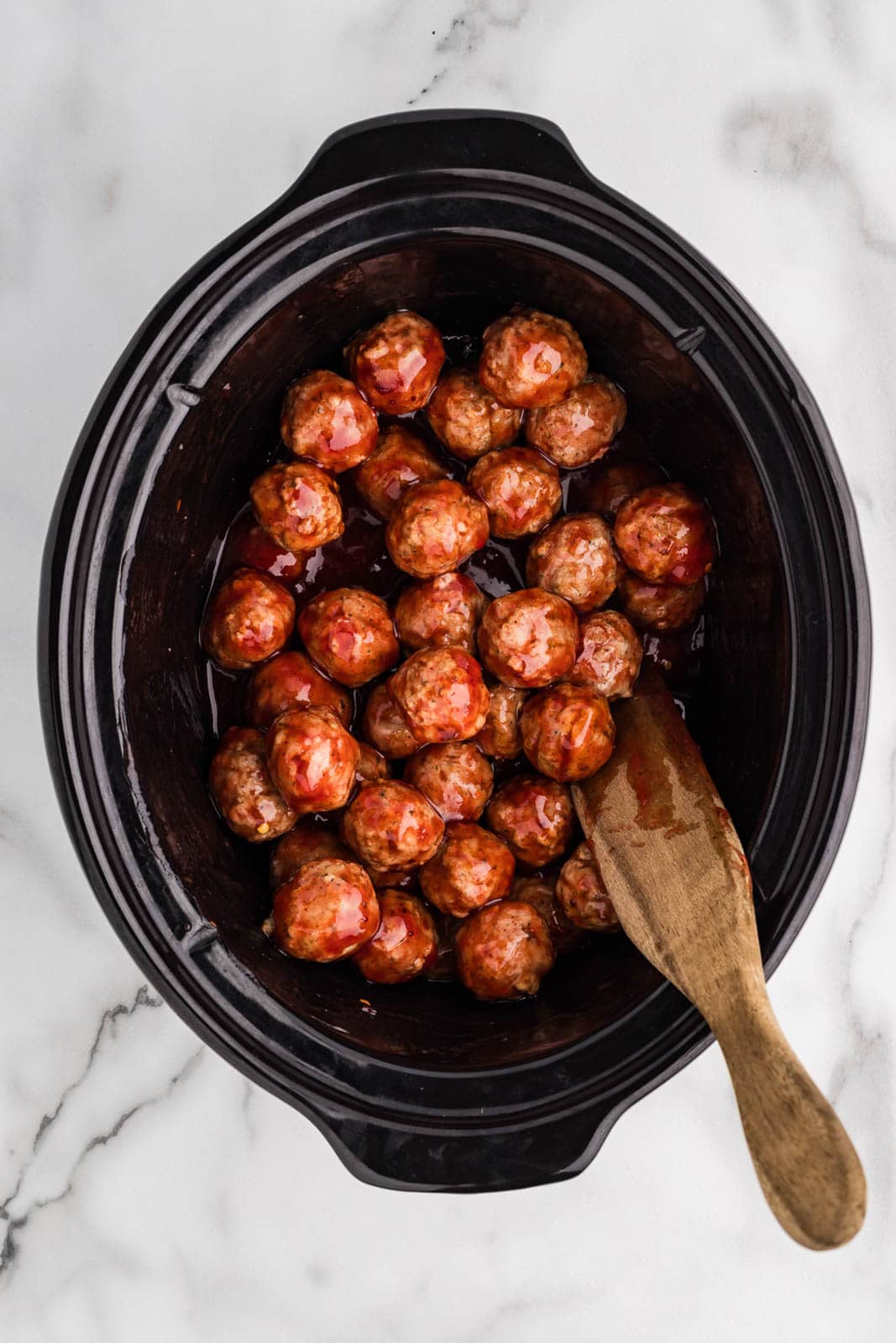 overhead view of black slow cooker containing cranberry meatballs with wooden spatula