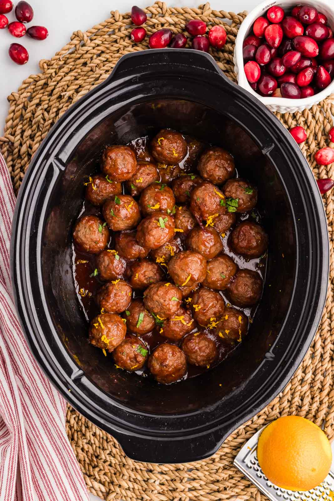overhead view of black slow cooker containing slow cooked cranberry meatballs on placemat and bowl of cranberries on side