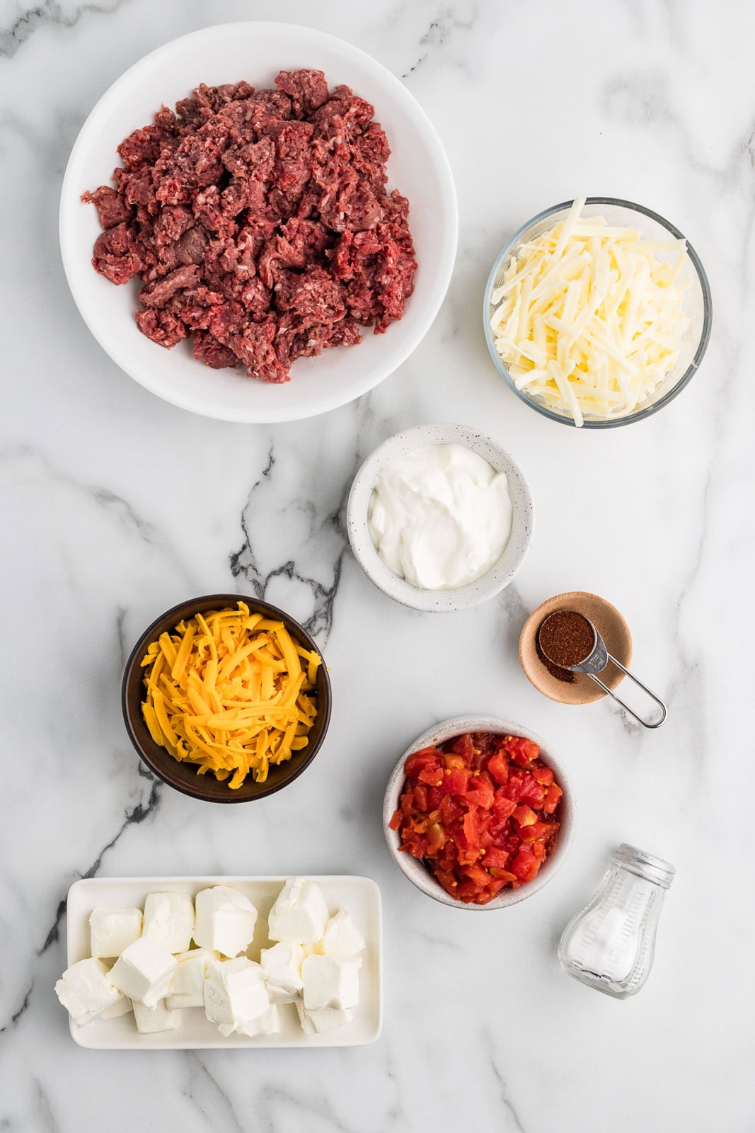 ingredients to include bowls of ground beef, Rotel, cream cheese, cheddar cheese, Monterey Jack cheese, sour cream, chili powder, and a salt shaker