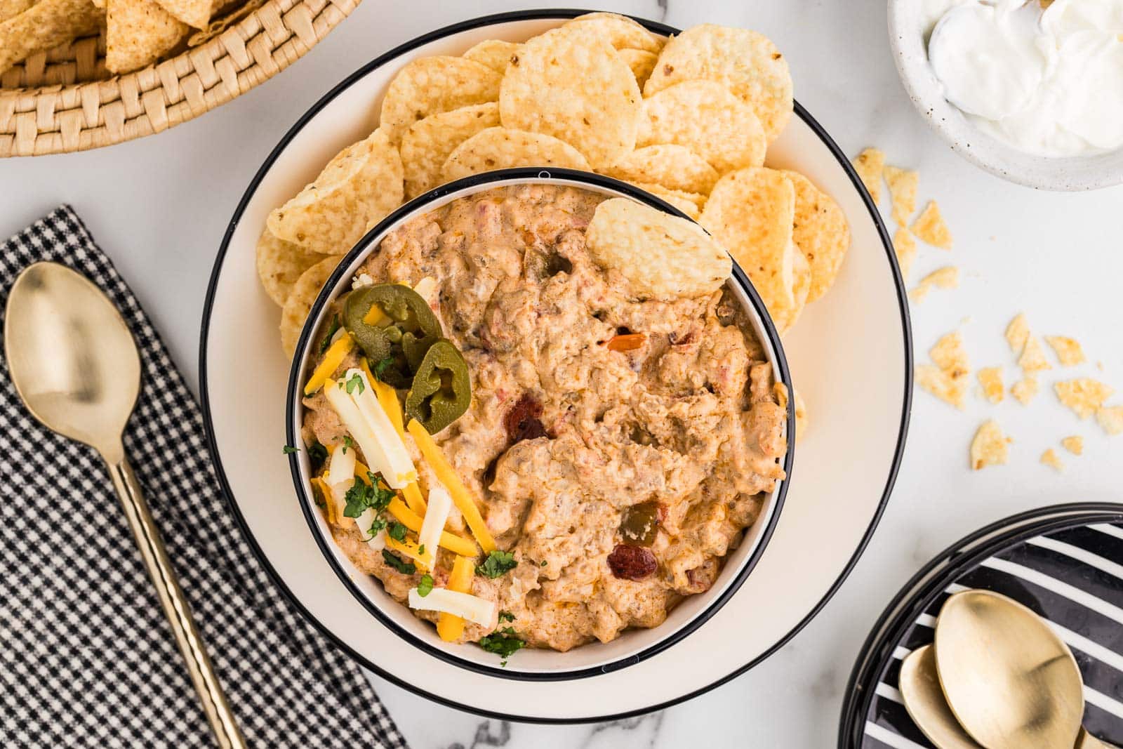 overhead view of dinner plate with chips and bowl of Rotel dip, surrounded by a gold spoons, plates and spoons, basket of chips and bowl of sour cream