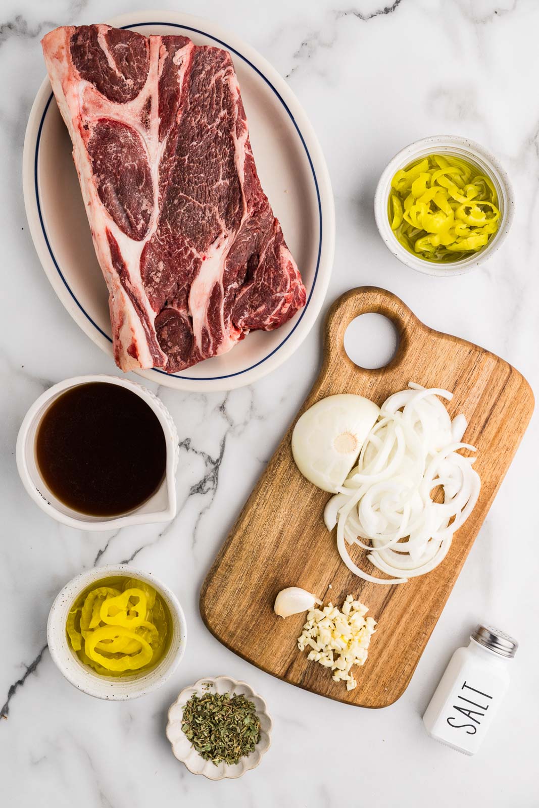 ingredients to include platter with chuck roast, wooden cutting board with slices onion and minced garlic, surrounded by bowls of sliced banana peppers, Italian seasoning, sliced pepperoncini, beef broth, and a salt shaker