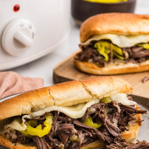 dinner plate with Italian beef sub with cheese, with cutting board holding another sub, slow cooker and bowl of banana peppers in background