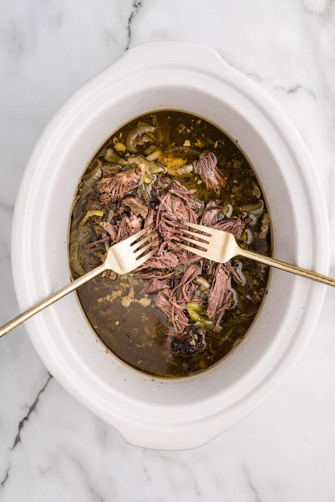 overhead view of white slow cooker with slow cooked Italian beef with two forks shredding the beef