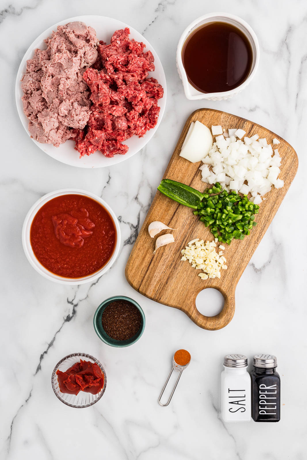 ingredients to include white plate of ground beef and ground turkey, bowl of beef broth, wooden cutting board with diced onion diced jalapenos, and minced garlic, salt and pepper shakers, teaspoon of smoked paprika, bowls of tomato paste, tomato sauce and bowl of chili seasoning