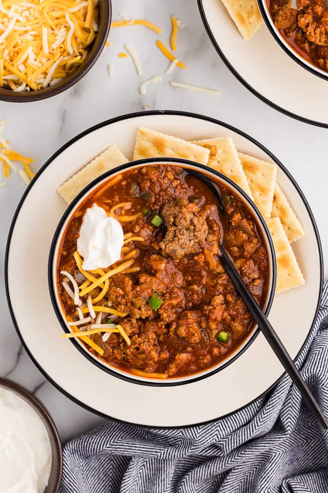 overhead view of white dinner plate with bowl of chili topped with cheese and sour cream, crackers on the side, with bowls of cheese and sour cream and another bowl of chili in background