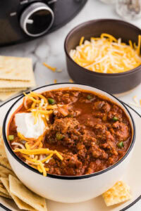 dinner plate with bowl of chili with sour cream and cheese on top, crackers on side, with bowl of cheese, crackers and black slow cooker in background