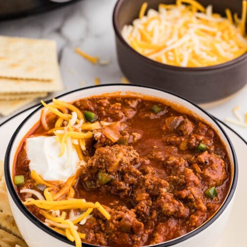 dinner plate with bowl of chili with sour cream and cheese on top, crackers on side, with bowl of cheese, crackers and black slow cooker in background