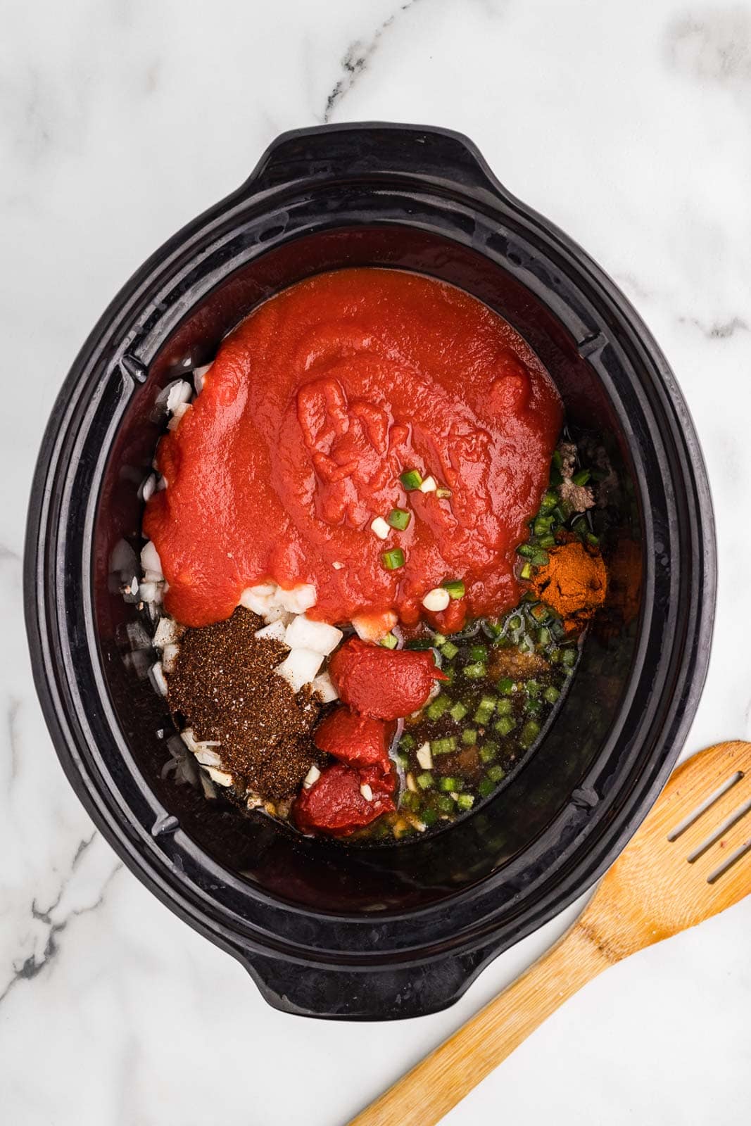 overhead view of black slow cooker with all ingredients with wooden spoon on side