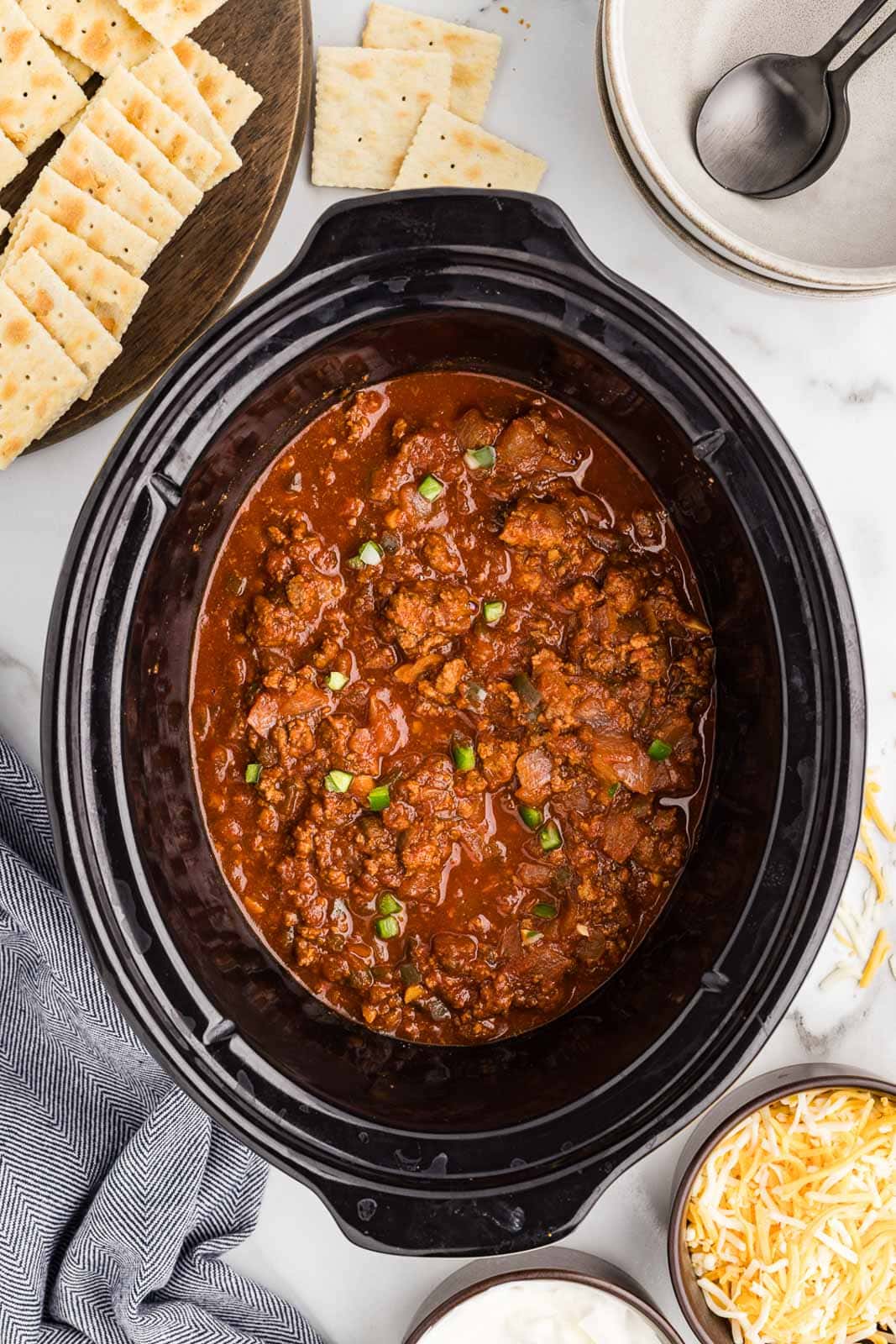 slow cooked chili recipe in black slow cooker, surrounded by crackers, bowls with spoons, cheese and sour cream