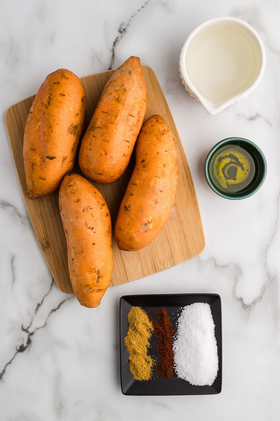 ingredients to contain four sweet potatoes on a wooden cutting board, surrounded by containers of water, olive oil, and flat dish with spices