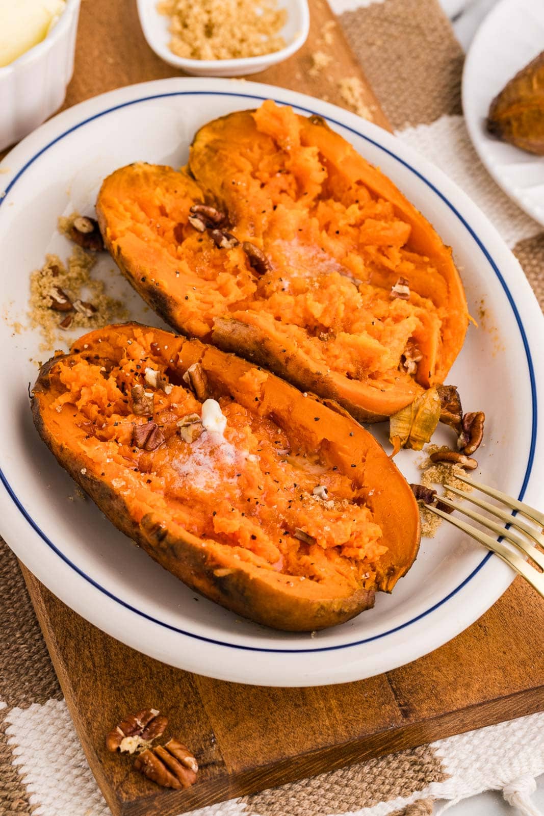 overhead view of white dinner plate on wooden cutting board containing two sweet potatoes that have been sliced open with butter on top and fork on the side