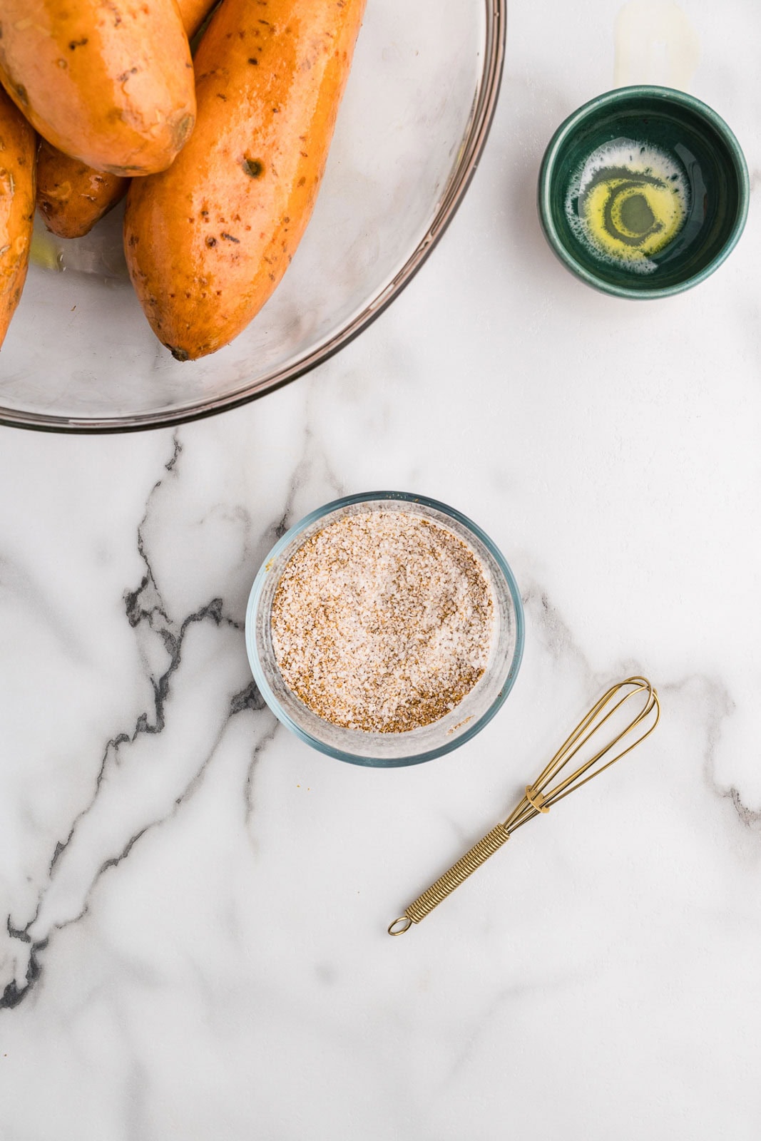 clear bowl of sweet potatoes, bowl of olive oil and glass bowl of all spices mixed together, with small whisk on side