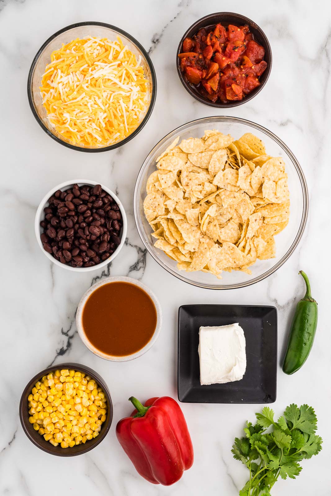 ingredients to include bowls of tortilla chips, black beans, shredded cheese, diced tomatoes, frozen corn, enchilada sauce, cream cheese, red bell pepper, jalapeno pepper, cilantro