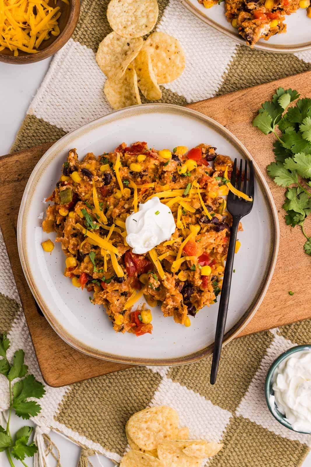 overhead view of dinner plate on wooden cutting board containing taco casserole with sour cream on top, fork on the side, with bowls of cheese and sour cream on side