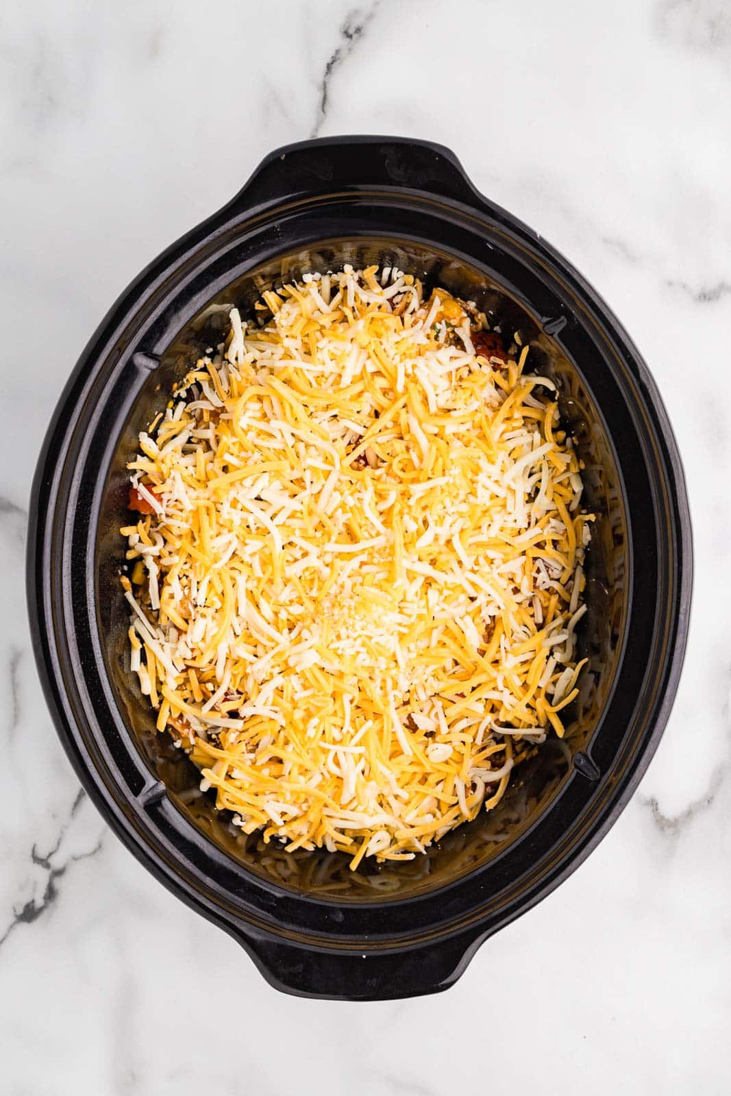 overhead view of black slow cooker with all ingredients and extra cheese on top