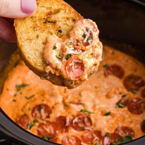 closeup of black slow cooker with pizza dip and hand holding bread that has been dipped in pizza dip