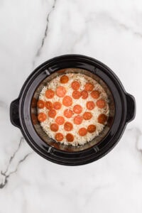 black slow cooker with pepperoni on top of cheese mixture
