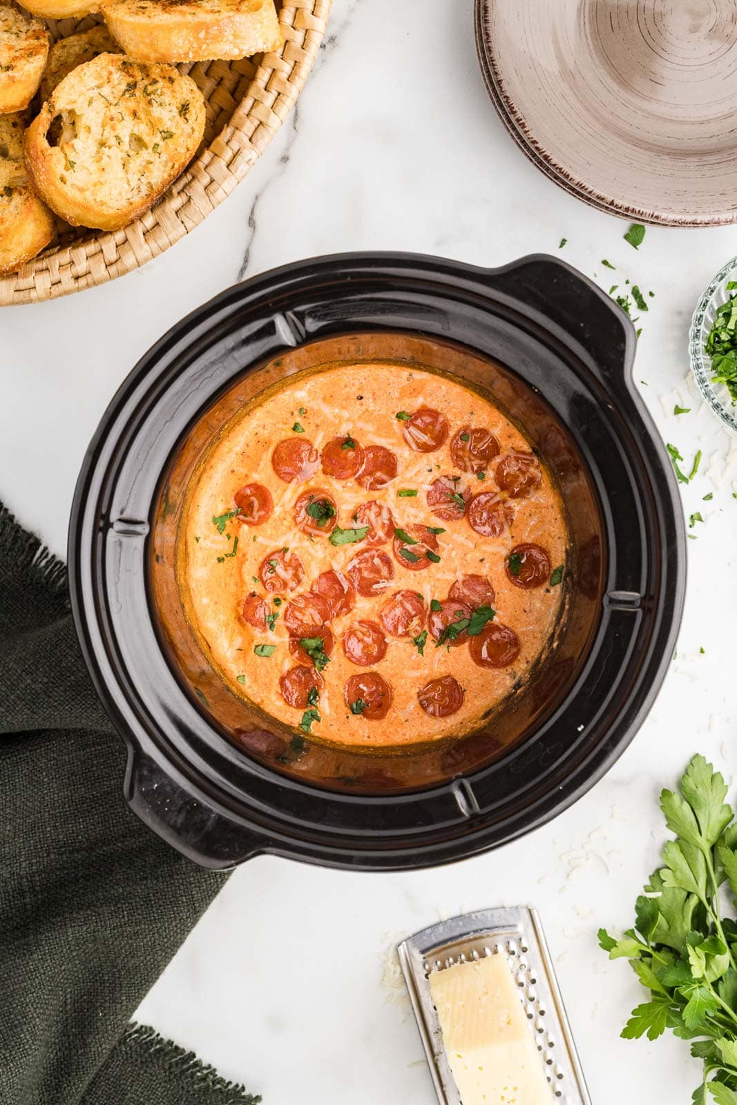 black slow cooker with pizza dip surrounded by basket of bread, Parmesan cheese and grater, and serving plate