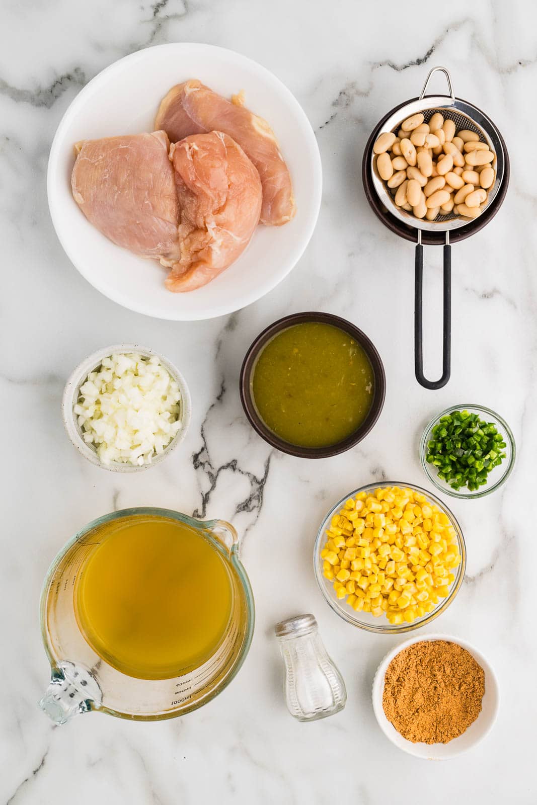 ingredients to include white plate of chicken breasts, bowl and strainer with white beans, black bowl of green enchilada sauce, bowls of diced jalapeno, frozen corn, taco seasoning, diced onion, shaker of salt, and glass measuring cup of chicken broth