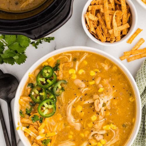 overhead view of white bowl of soup with sliced jalapeno, surrounded by bowl of tortilla strips, black spoons and black slow cooker in background