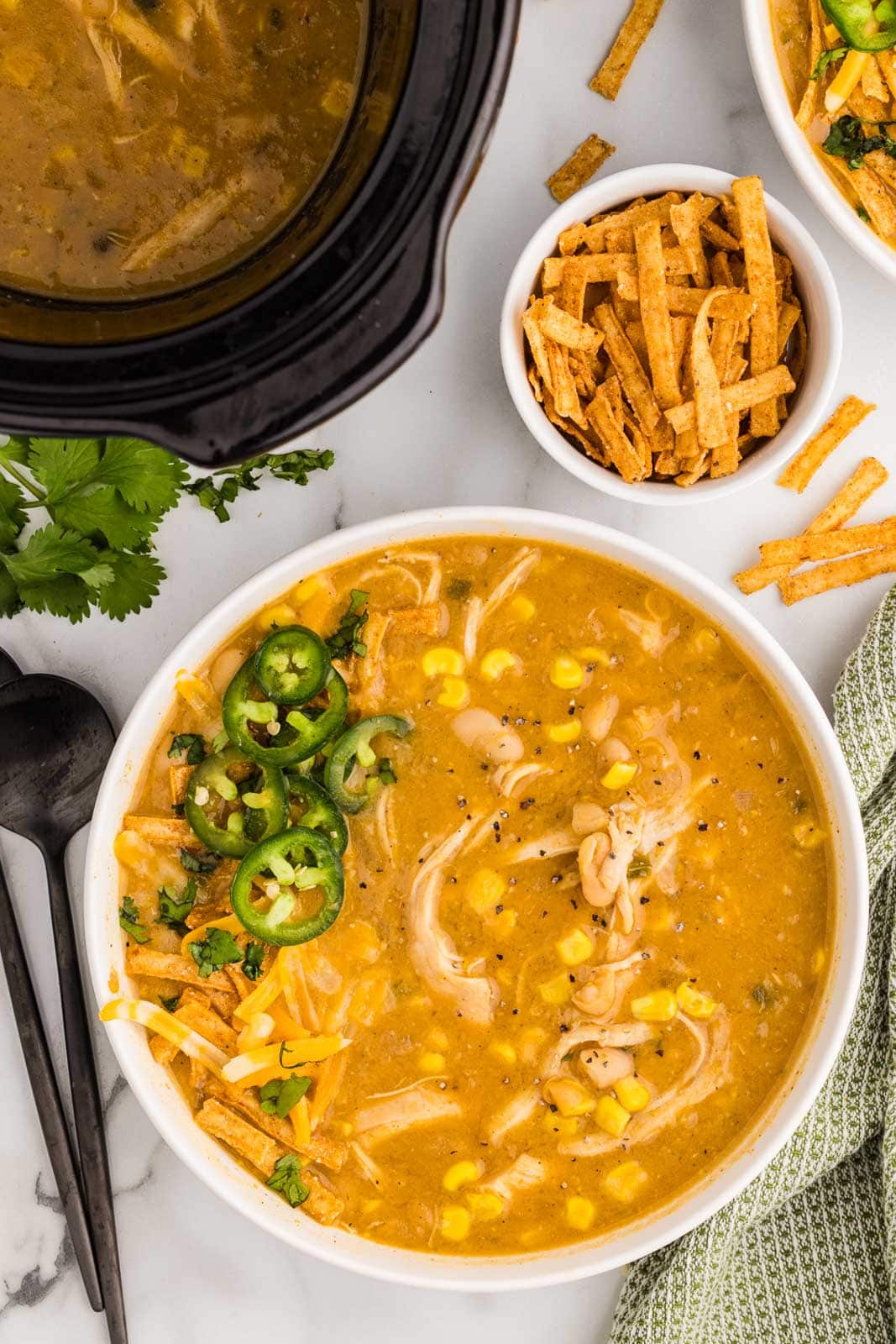 overhead view of white bowl of soup with sliced jalapeno, surrounded by bowl of tortilla strips, black spoons and black slow cooker in background