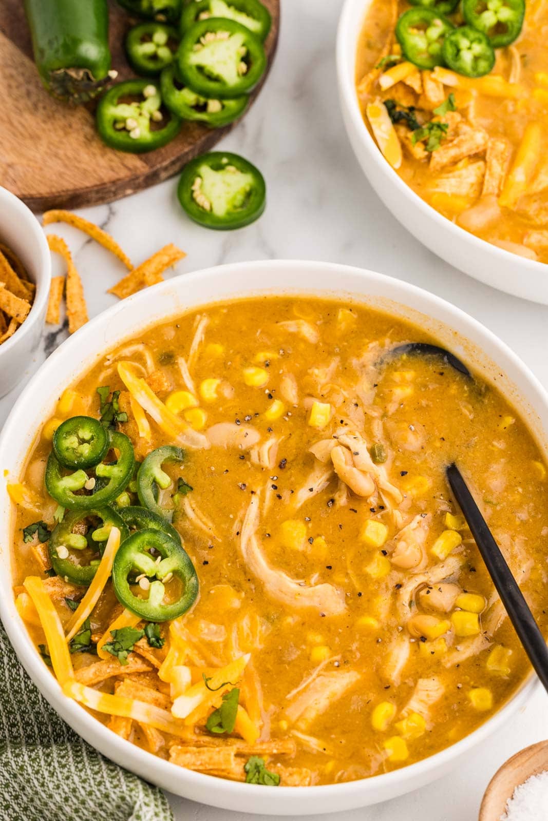 overhead view of white bowl of soup with black spoon inserted, surrounded by another bowl of soup and wooden cutting board with whole jalapeno and jalapeno slices