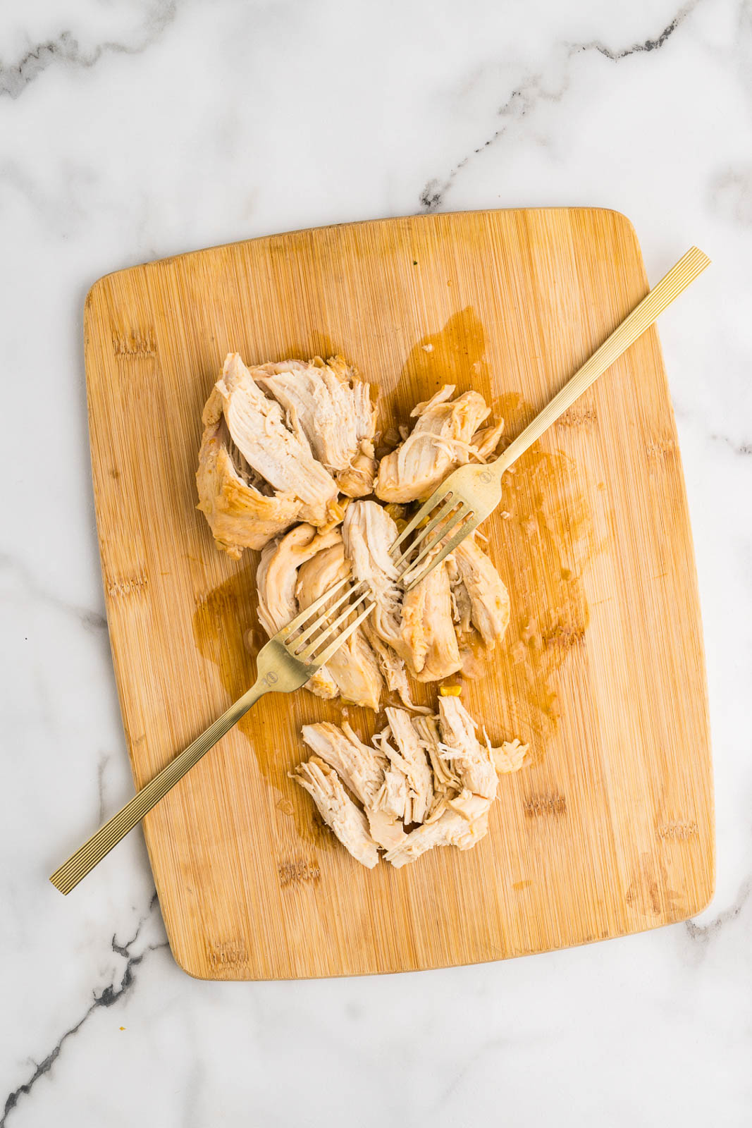 wooden cutting board with chicken pieces being shredded with two gold forks