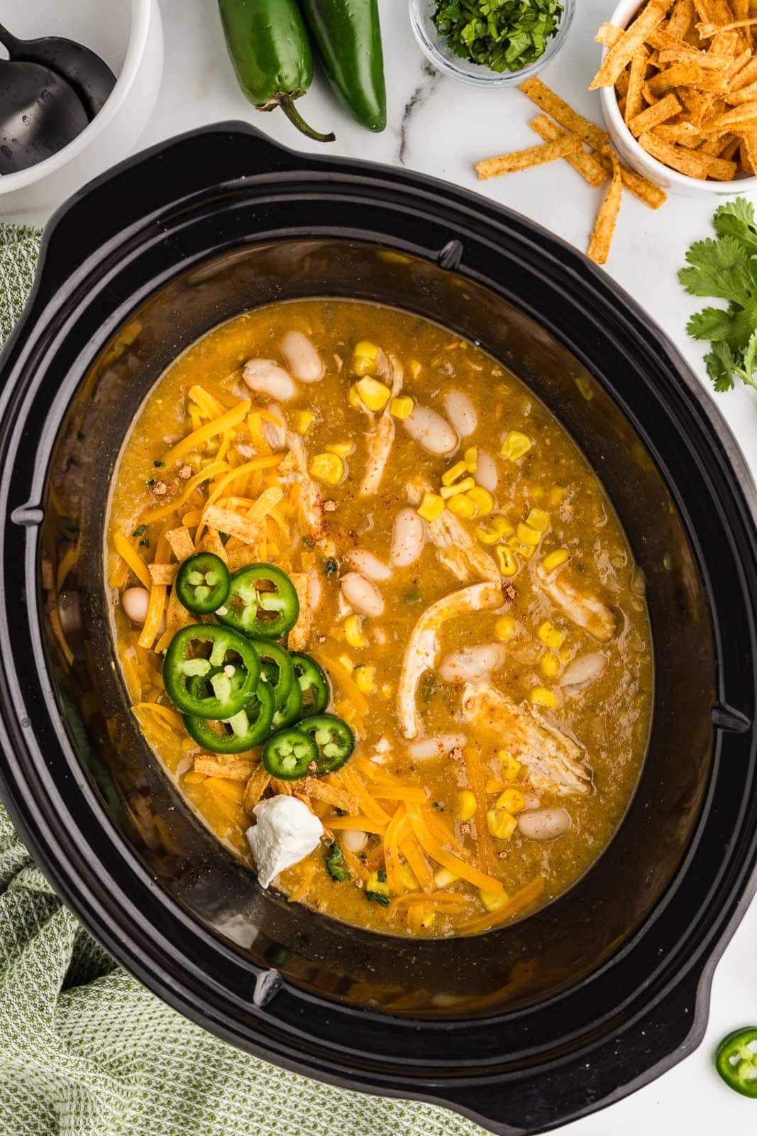 overhead view of black slow cooker containing soup with sliced jalapeno peppers on top, surrounded by bowls of tortilla strips, diced jalapeno, avocado, and 2 whole jalapeno
