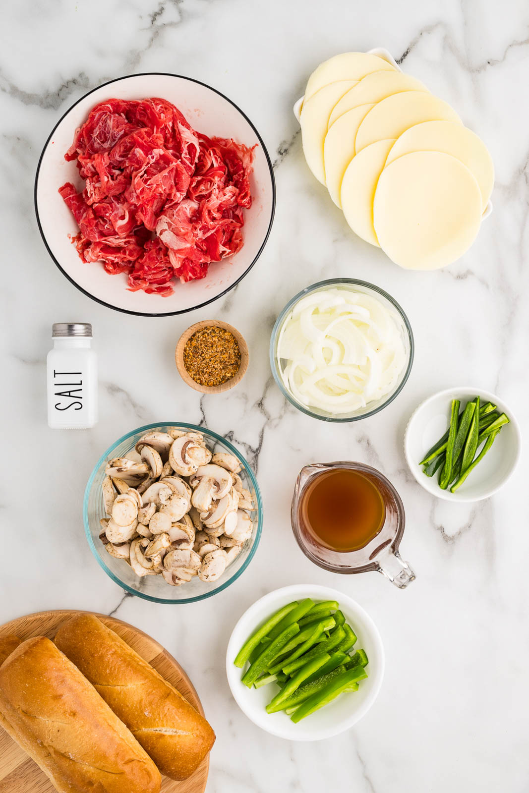 ingredients to include bowls of shaved beef steak surrounded by sliced onion, green bell pepper, mushrooms, jalapeno, salt shaker, steak seasoning, measuring cup of beef broth, sliced provolone cheese and wooden cutting board with hoagie rolls