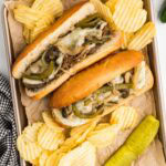 overhead view of parchment paper lined baking tin with two hoagies filled with beef steak mixture with melted cheese with potato chips and dill pickle on side