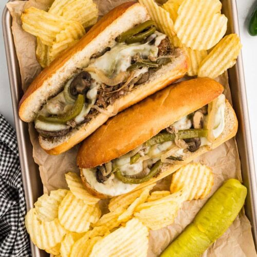 overhead view of parchment paper lined baking tin with two hoagies filled with beef steak mixture with melted cheese with potato chips and dill pickle on side