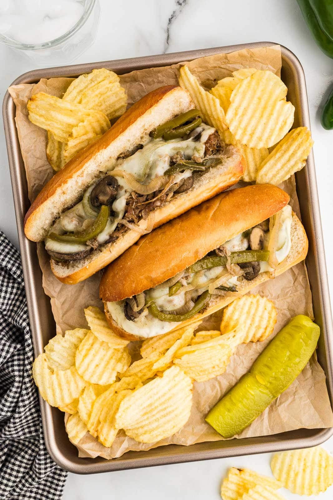 overhead view of parchment paper lined baking tin with two hoagies filled with beef steak mixture with melted cheese with potato chips and dill pickle on side
