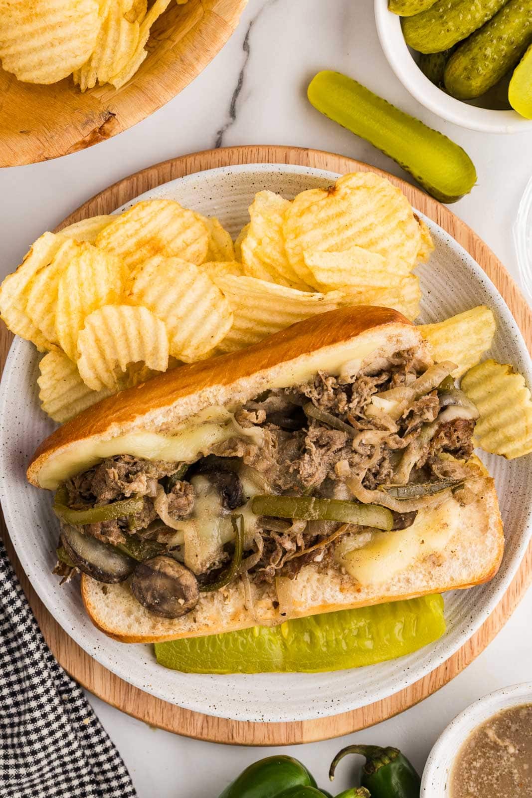 overhead view of white dinner plate on wooden cutting board with hoagie filled with beef steak mixture with melted cheese, surrounded by potato chips and dill pickle, with bowl of dill pickles and potatoes chips in background