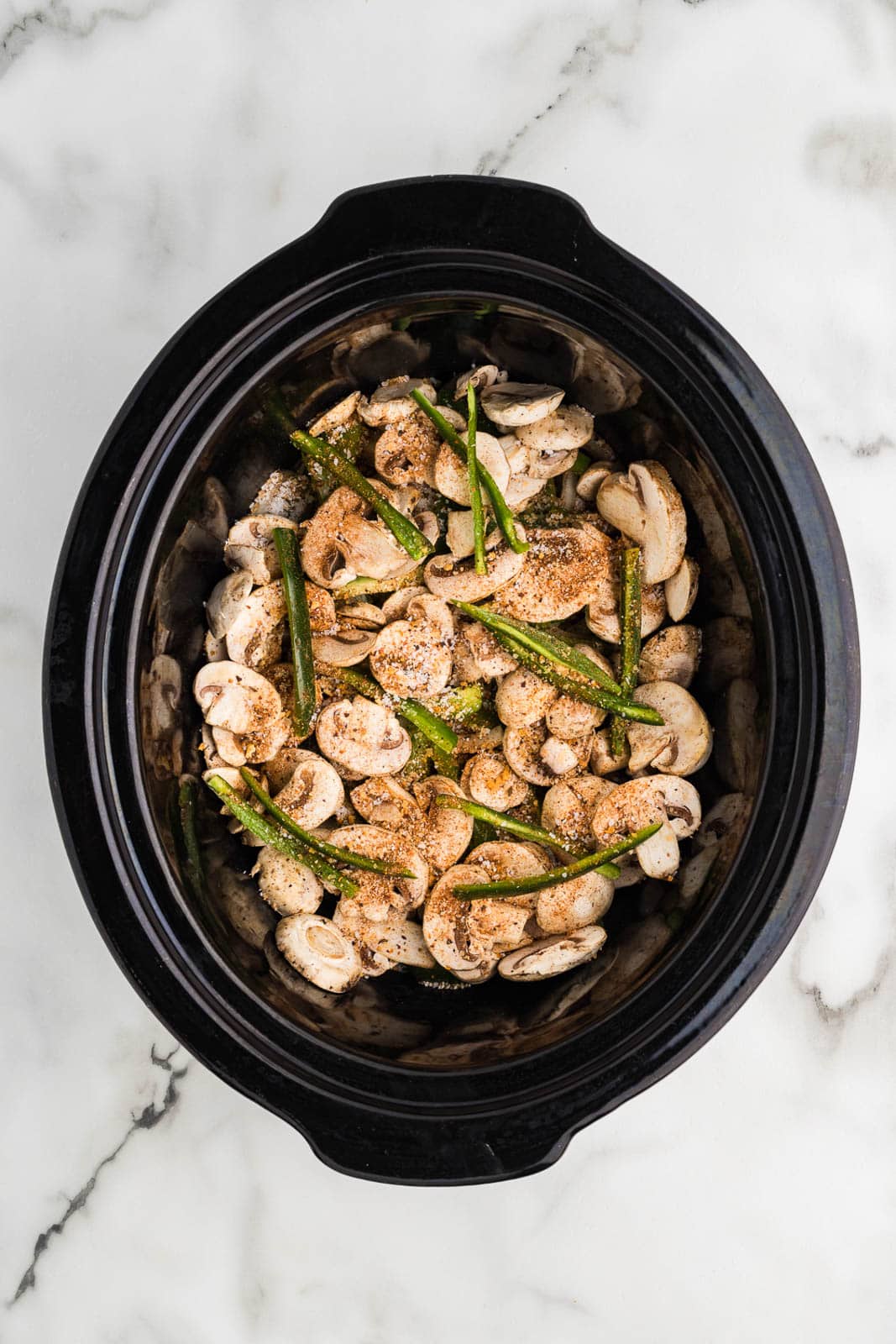 overhead view of black slow cooker with beef, mushrooms, peppers and onions with seasoning on top