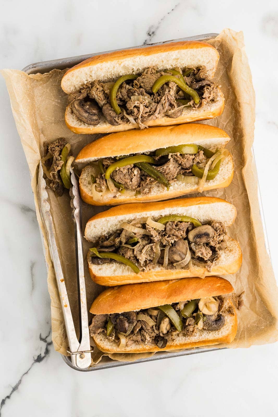 overhead view of parchment paper lined baking tin with four hoagies filled with beef steak mixture with metal tongs on side