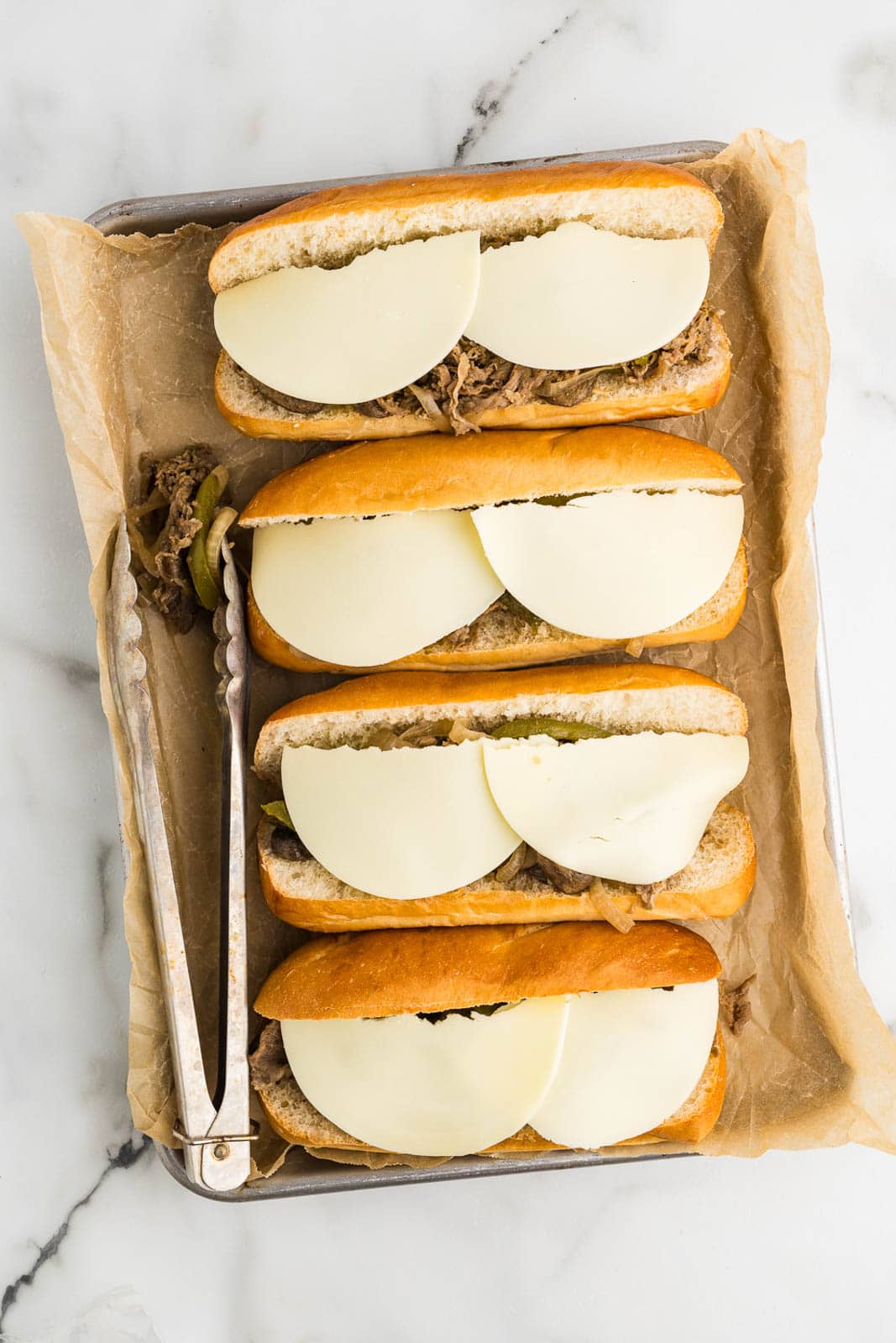 overhead view of parchment paper lined baking tin with four hoagies filled with beef steak mixture topped with cheese slices, with metal tongs on side