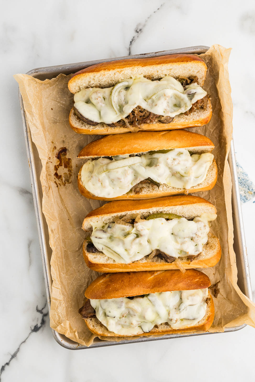 overhead view of parchment paper lined baking tin with four hoagies filled with beef steak mixture topped with melted cheese slices
