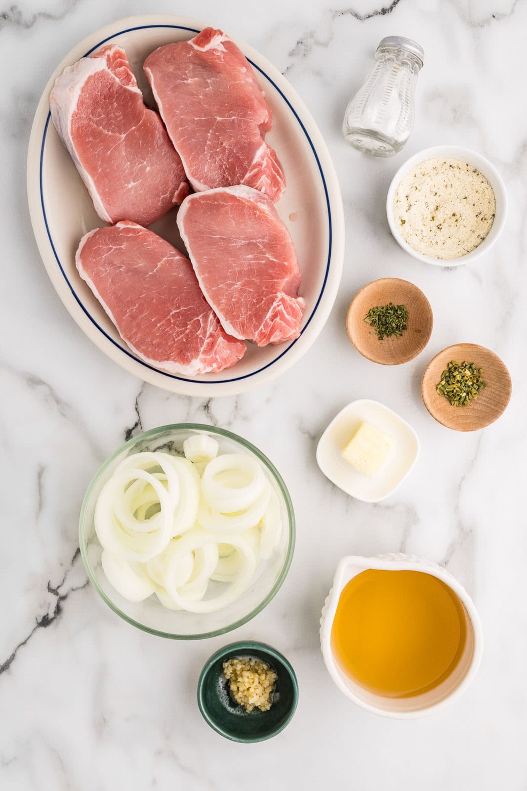 ingredients to include large platter with 4 boneless pork chops, surrounded by containers of sliced onion, ranch seasoning, chicken broth, butter, minced garlic, parsley, chives and salt shaker