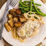white dinner plate containing one slow cooker pork chops with onions, green beans and potato wedges with dinner knife and fork on top of a wooden cutting board